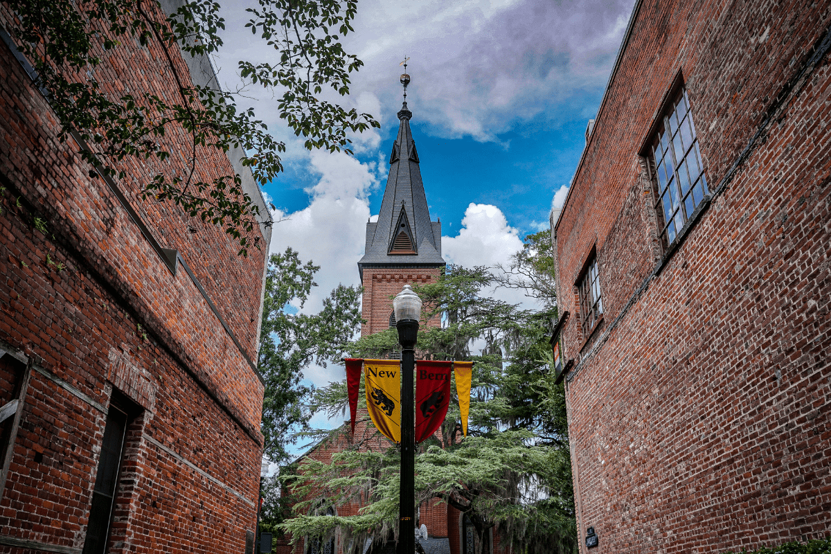 A tall brick church steeple with a clock is framed by two brick buildings, with a lamppost and colorful banners in the foreground under a partly cloudy sky.