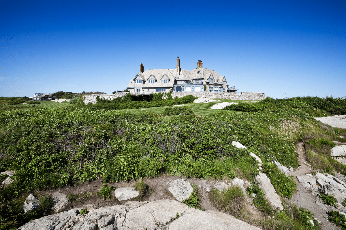 A large stone mansion with multiple chimneys sits atop a grassy hill, surrounded by lush greenery and rocks, under a clear blue sky.