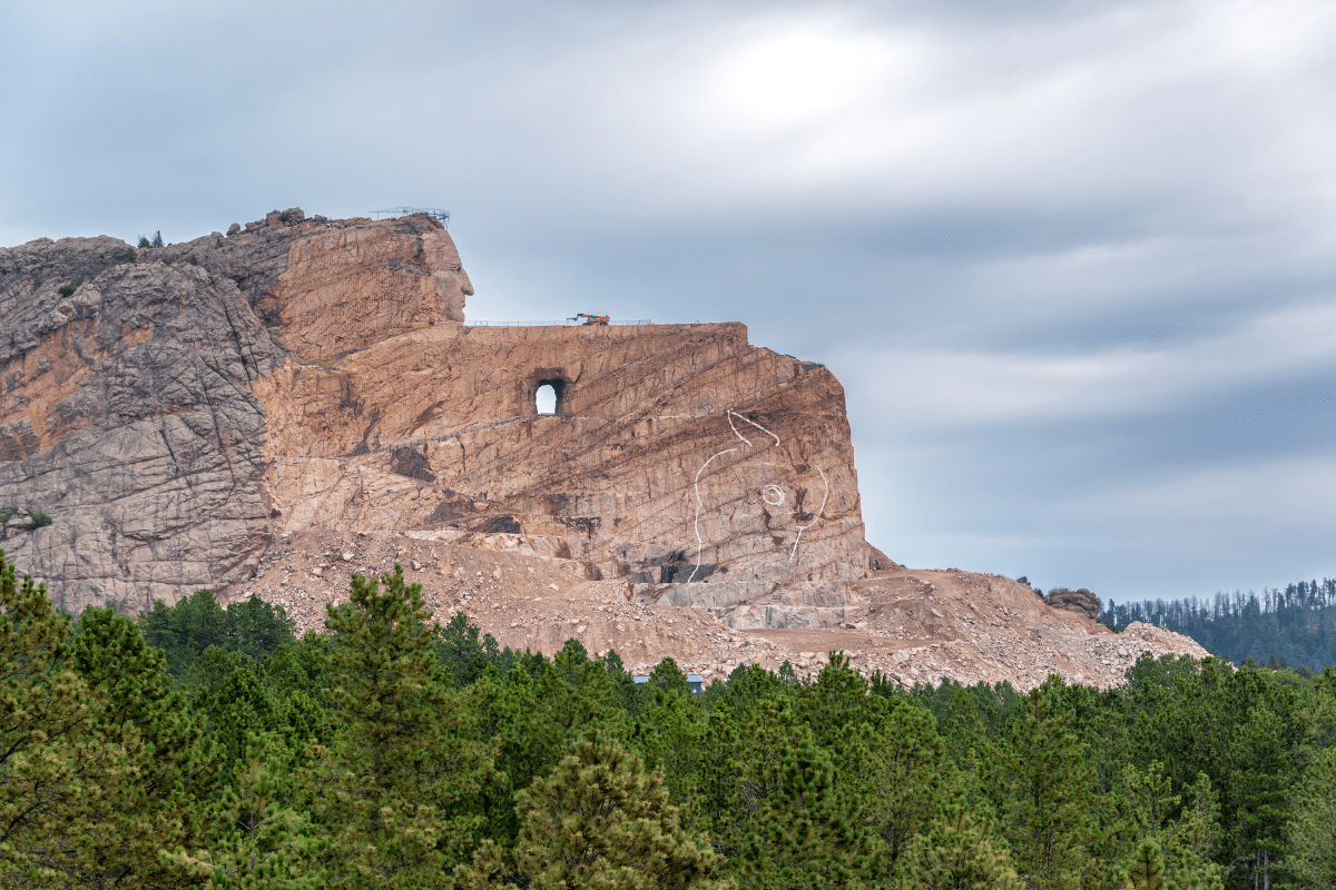 The image shows the partially completed Crazy Horse Memorial, a massive mountain carving depicting the Lakota leader, with a prominent hole where the horse's head will be, surrounded by green forest.
