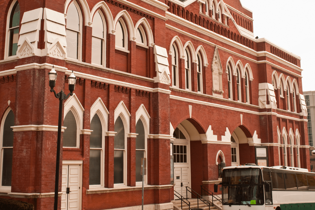 A historic red brick building with arched windows and white trim stands beside a parked tour bus, featuring ornate architectural details and multiple stories.