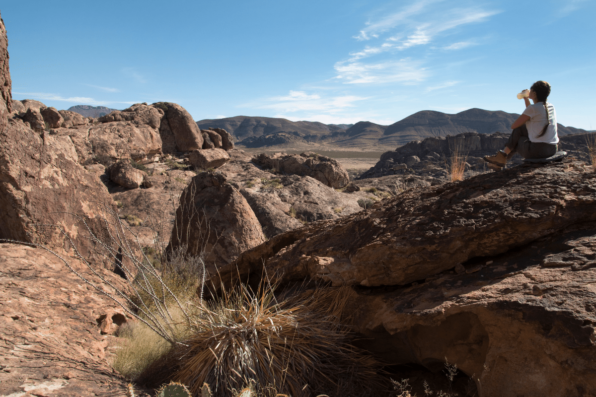 A person sits on a large rock in a desert landscape, drinking from a bottle, surrounded by rugged terrain and distant mountains under a clear blue sky.