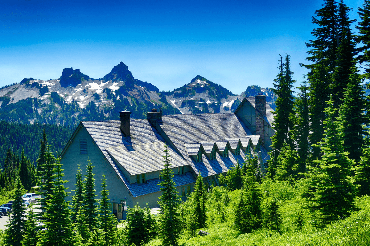 A large lodge with a steep roof is nestled among lush green trees, with rugged, snow-dusted mountains in the background under a clear blue sky.