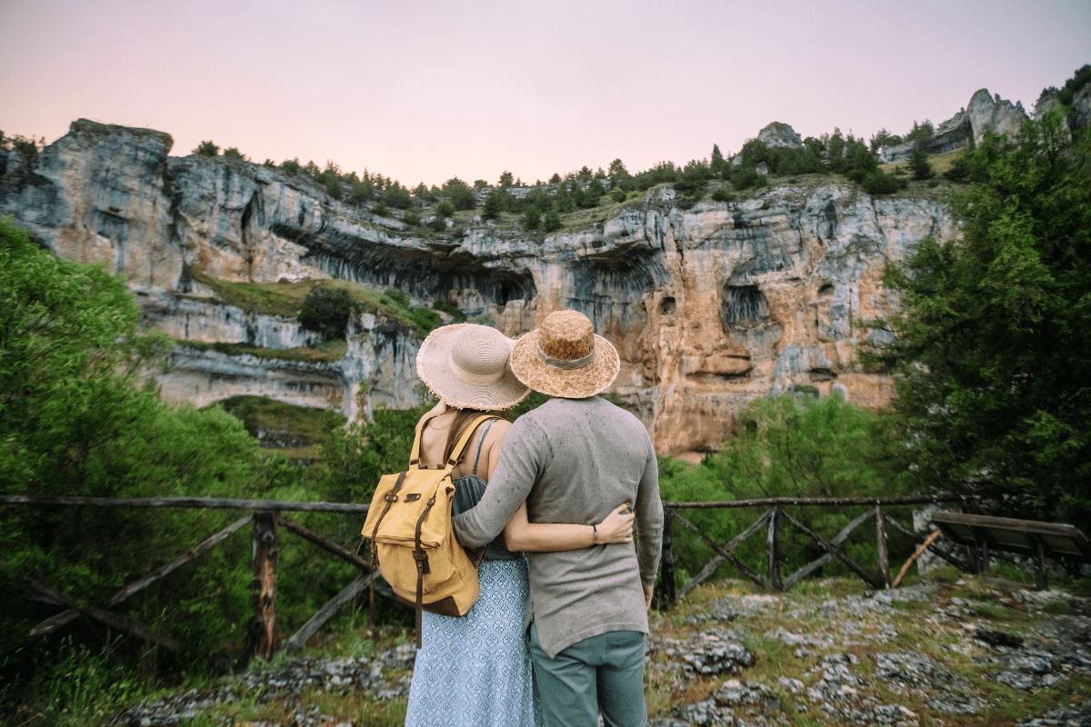 A couple wearing hats and casual clothing stands arm in arm, facing a scenic view of rocky cliffs and greenery under a cloudy sky.