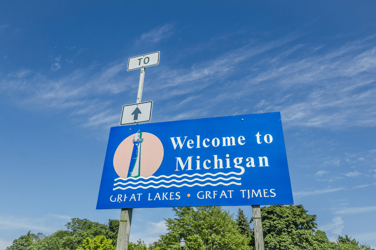 A blue sign with a lighthouse illustration reads "Welcome to Michigan, Great Lakes, Great Times" against a clear sky, with trees in the background and a directional sign above.