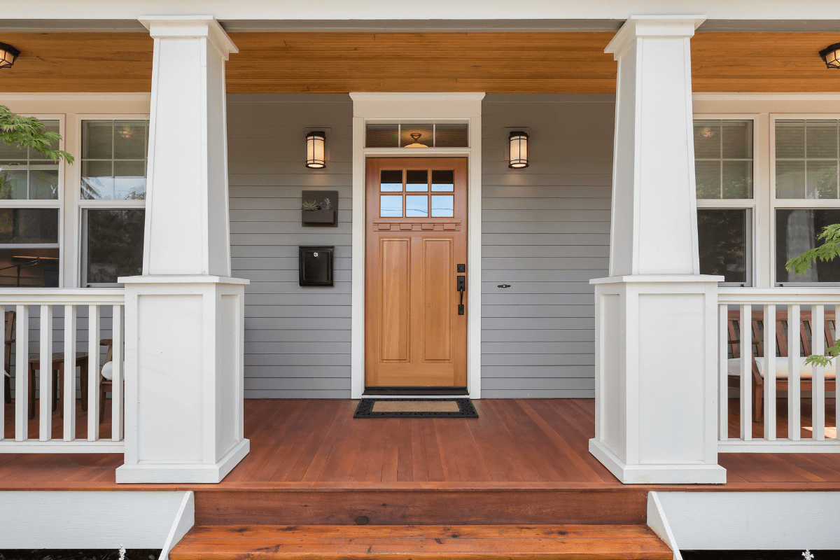 A welcoming front porch features a wooden door with a glass panel, flanked by two windows, white columns, and a wooden floor, creating a warm and inviting entrance.