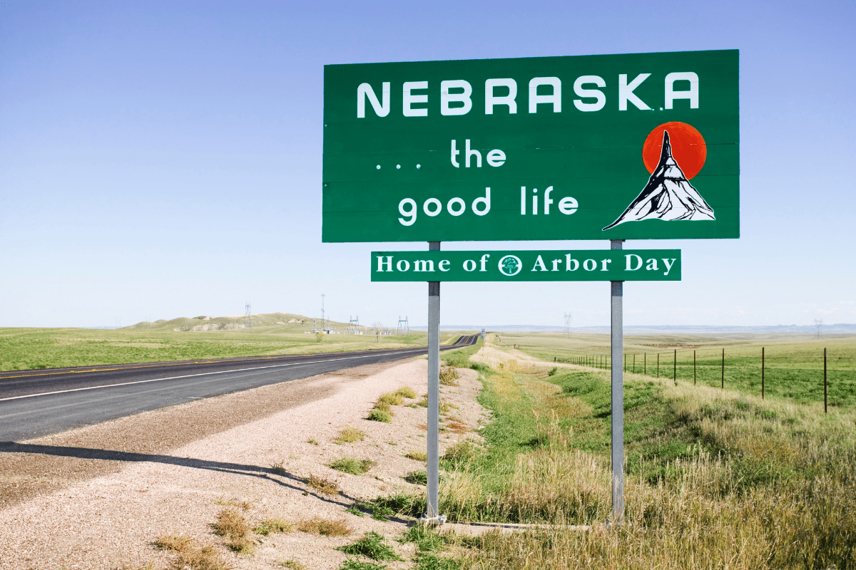 A roadside sign in a grassy landscape reads "Nebraska... the good life" with a sun and mountain graphic, and "Home of Arbor Day" below.