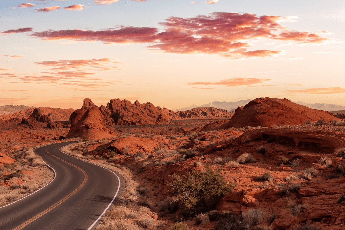 A winding road curves through a desert landscape with red rock formations and sparse vegetation under a sky filled with scattered pink clouds at sunset.