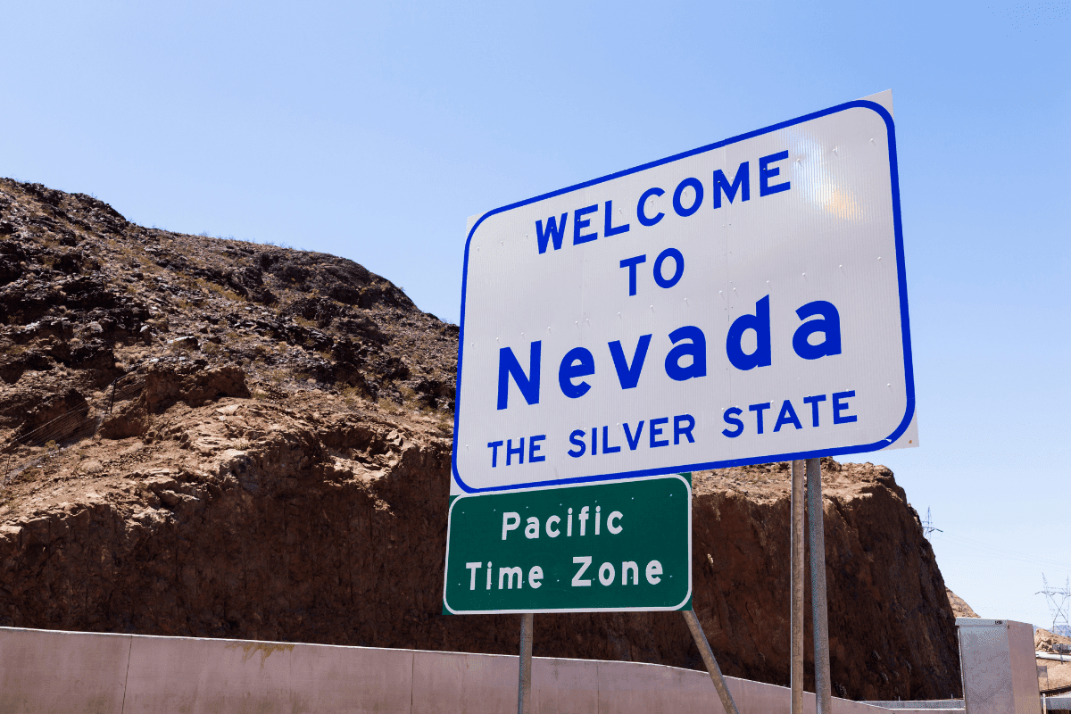 A "Welcome to Nevada, The Silver State" sign stands against a rocky landscape, with an additional sign indicating the Pacific Time Zone beneath it.
