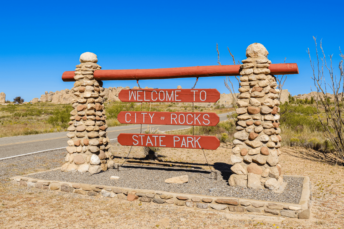 A stone and wood sign reads "Welcome to City of Rocks State Park" against a backdrop of desert landscape and clear blue sky.