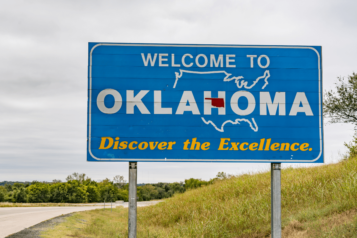 A blue roadside sign reads "Welcome to Oklahoma" with a map outline and the phrase "Discover the Excellence," set against a grassy landscape and overcast sky.
