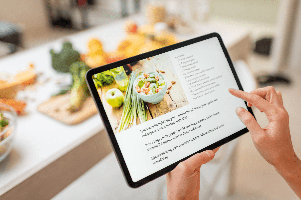 A person holds a tablet displaying a salad recipe, with a kitchen table in the background featuring fresh vegetables and ingredients.
