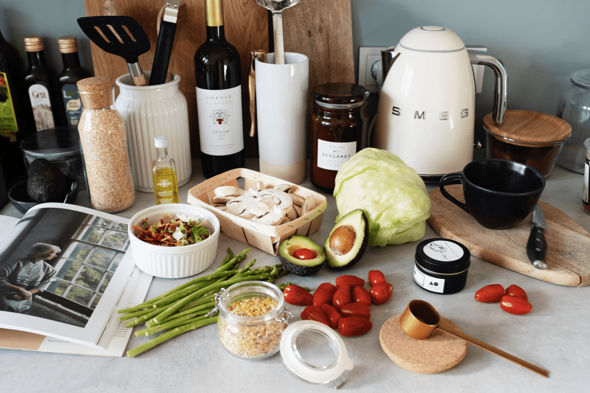 A kitchen counter displays various cooking ingredients, including fresh vegetables, wine bottles, a cookbook, and a Smeg kettle, suggesting a meal preparation scene.