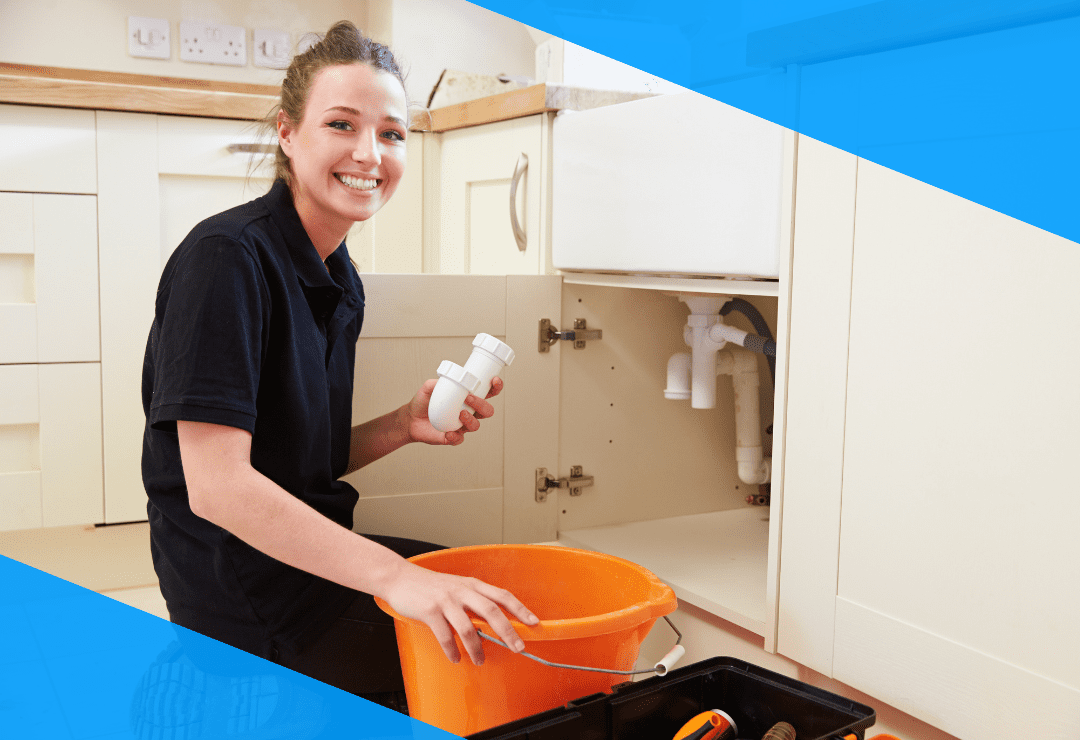 Person smiling while fixing plumbing under a kitchen sink, holding a pipe piece, with tools nearby.