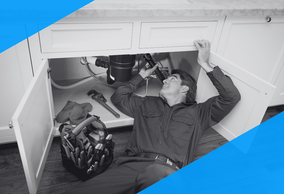 A plumber lies under a kitchen sink, repairing pipes with tools nearby.
