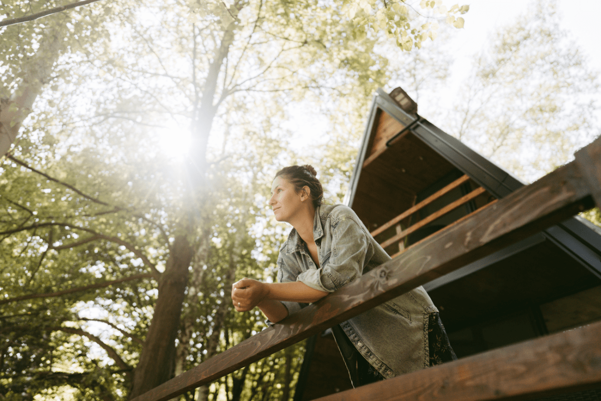 A person in a denim jacket leans on a wooden railing, gazing thoughtfully at the sunlit forest surrounding a modern cabin.