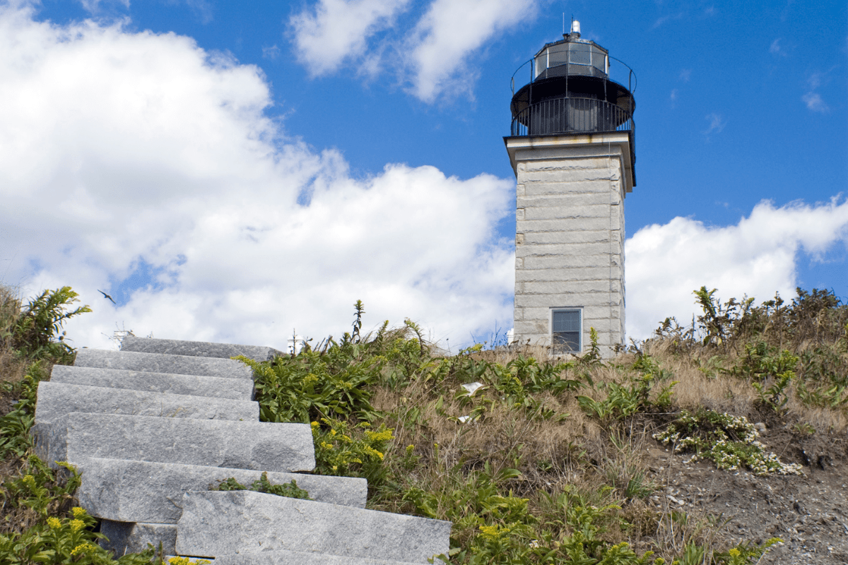 A stone lighthouse stands under a blue sky with scattered clouds, surrounded by grassy terrain and wild vegetation, with stone steps leading up to it.