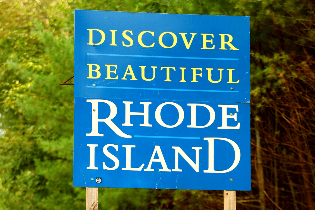 A blue sign with yellow and white text reads "Discover Beautiful Rhode Island" against a backdrop of green foliage.