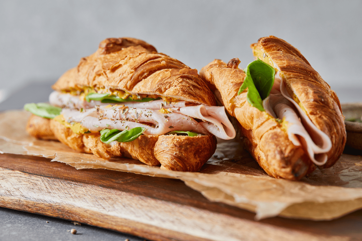A croissant sandwich filled with sliced turkey, fresh spinach, and a spread, placed on parchment paper over a wooden board.
