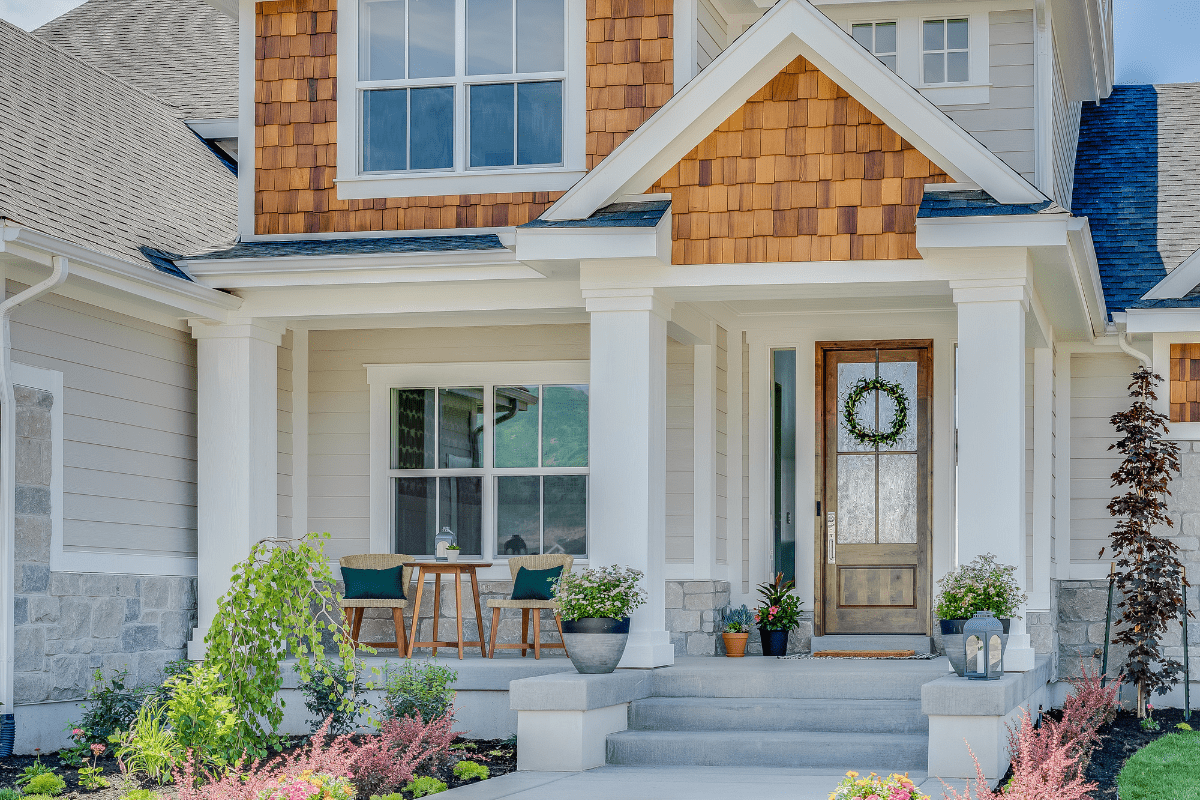 A modern suburban house features a welcoming front porch with a wooden door, a wreath, and a small seating area surrounded by neatly landscaped greenery.