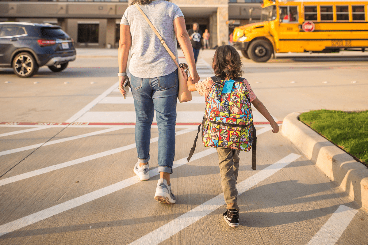 A woman and a child with a colorful backpack walk hand-in-hand across a parking lot towards a yellow school bus.