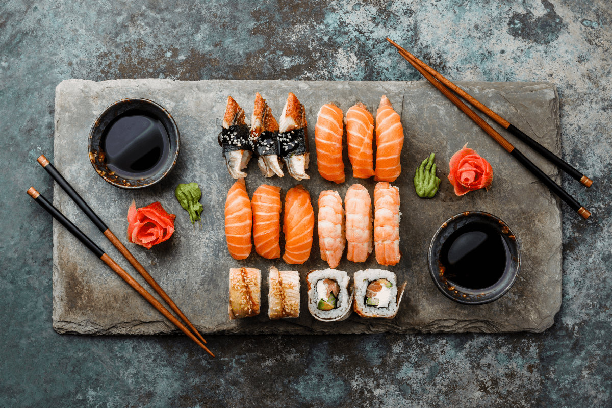 A slate platter displays an assortment of sushi, including nigiri and rolls, accompanied by soy sauce, wasabi, and ginger, with chopsticks placed on either side.