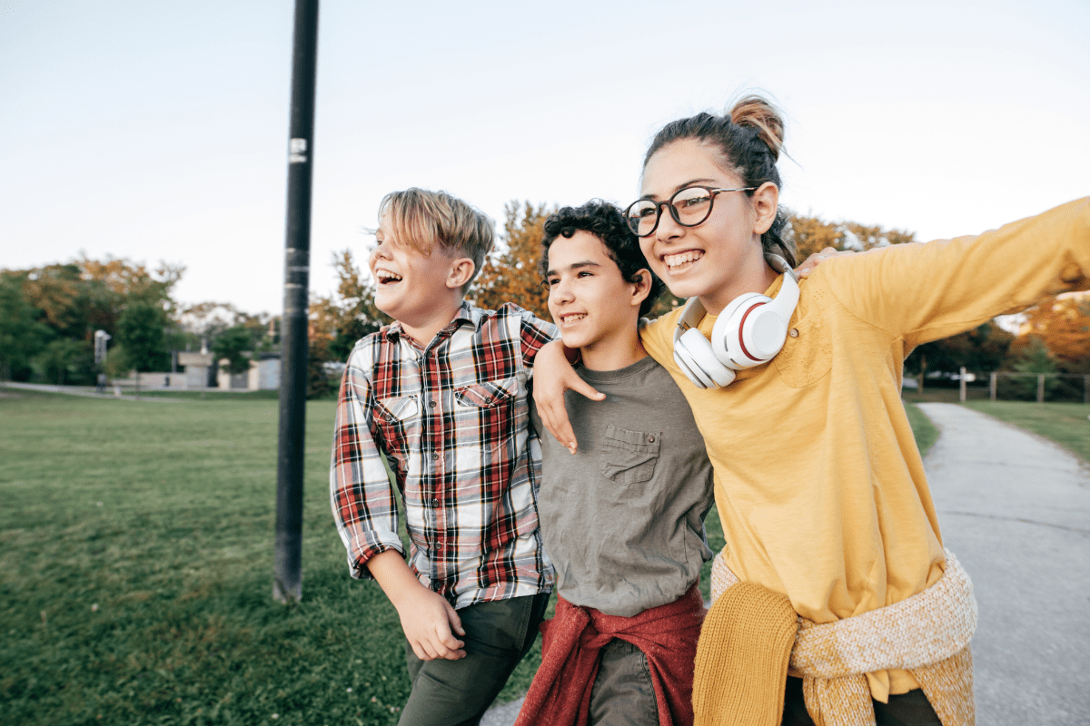 Three smiling children walk together on a path in a park, with one wearing headphones and a yellow sweater, enjoying a sunny day.