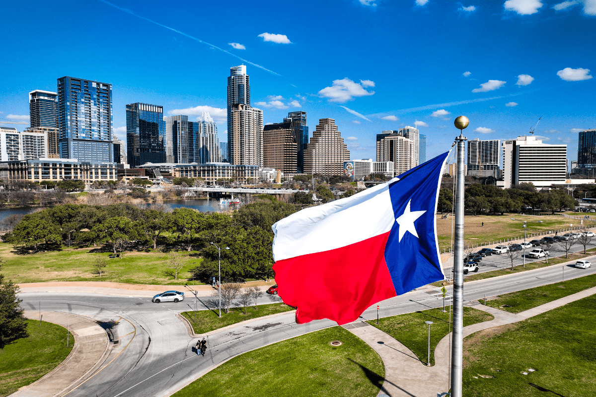 A large Texas flag waves prominently in the foreground with a backdrop of a modern city skyline under a clear blue sky.