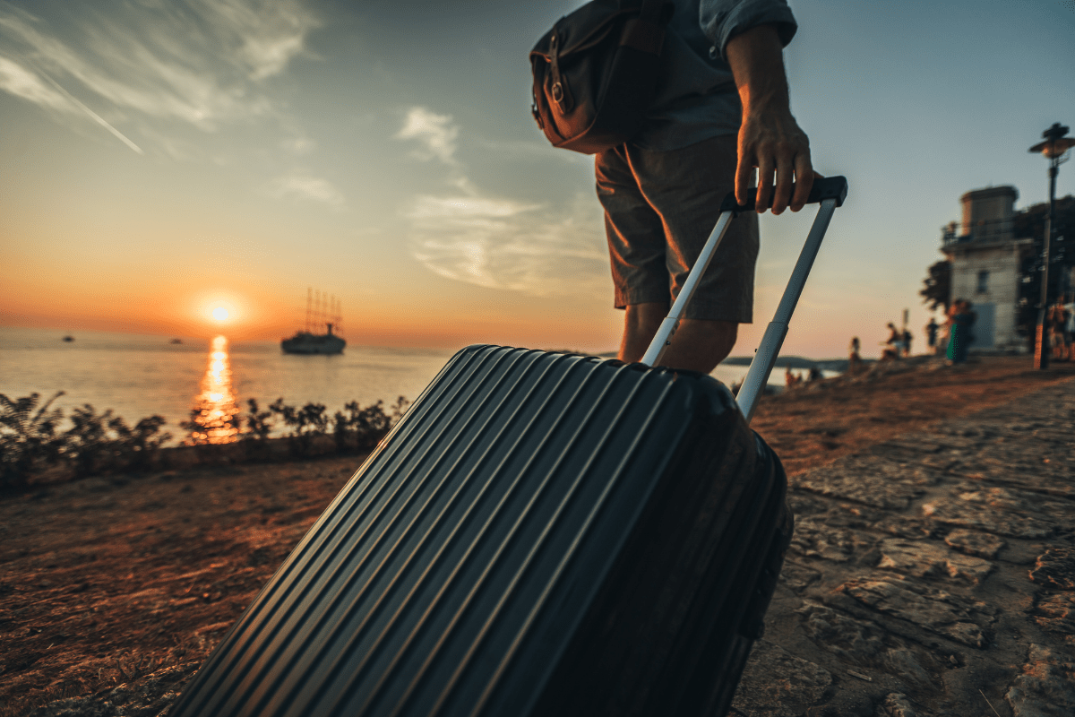 A person pulls a suitcase along a cobblestone path at sunset, with a ship visible on the horizon and a lighthouse nearby.