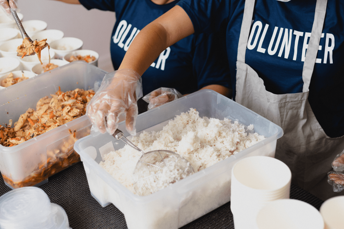 Volunteers wearing gloves and "VOLUNTEER" shirts prepare and serve rice and food into containers, likely for a community or charity event.