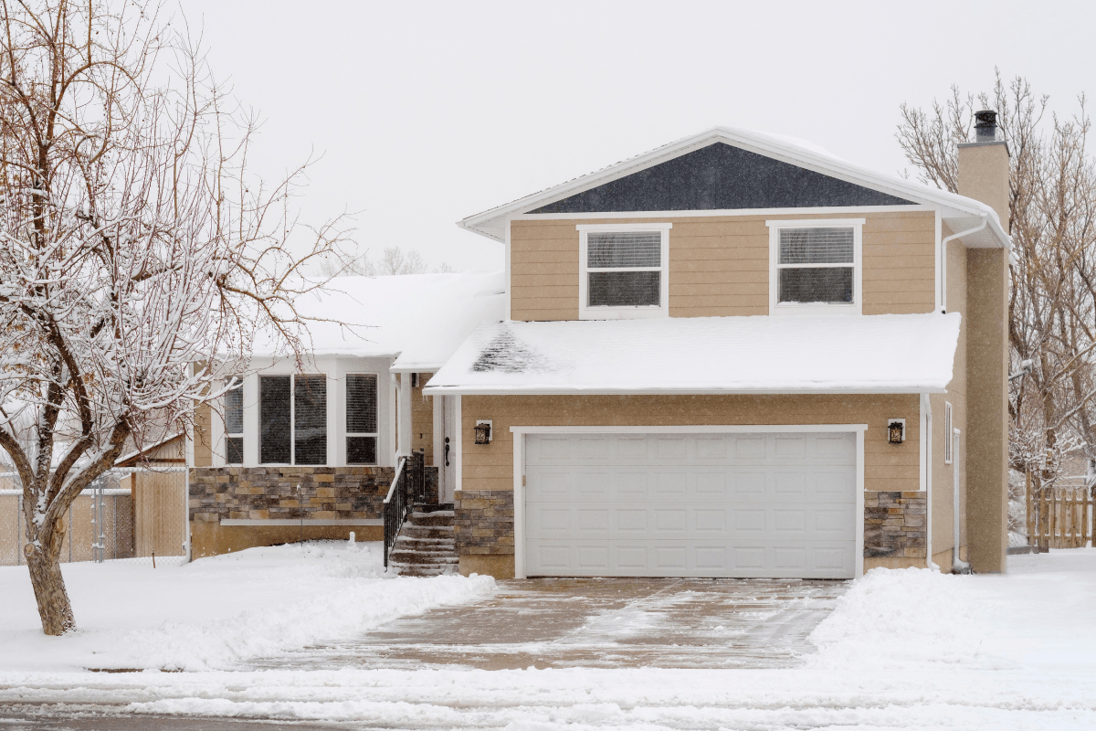 A two-story house with a beige exterior and a large garage is surrounded by snow-covered trees and ground, creating a serene winter scene.