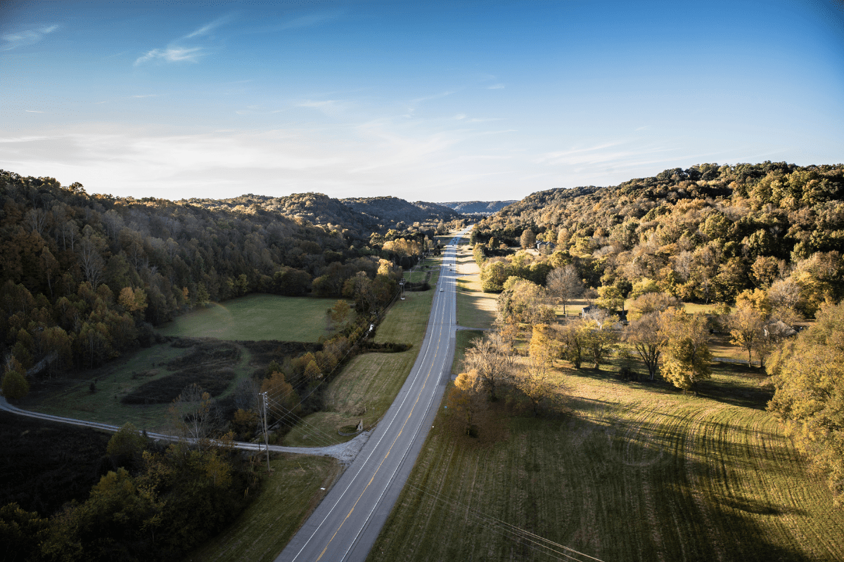 Aerial view of a scenic rural road winding through a lush, hilly landscape with trees displaying autumn colors under a clear blue sky.