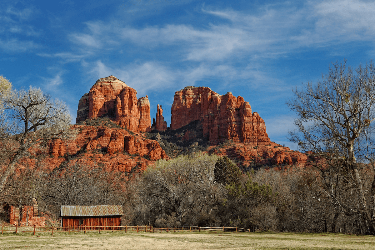 A scenic view of Sedona's red rock formations under a blue sky, with a rustic barn and leafless trees in the foreground.