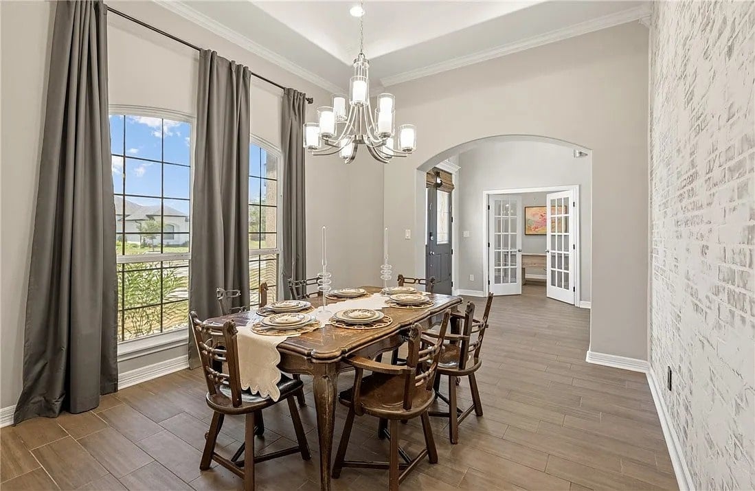 Dining room with wooden table, chairs, chandelier, large windows, and view into adjacent room.