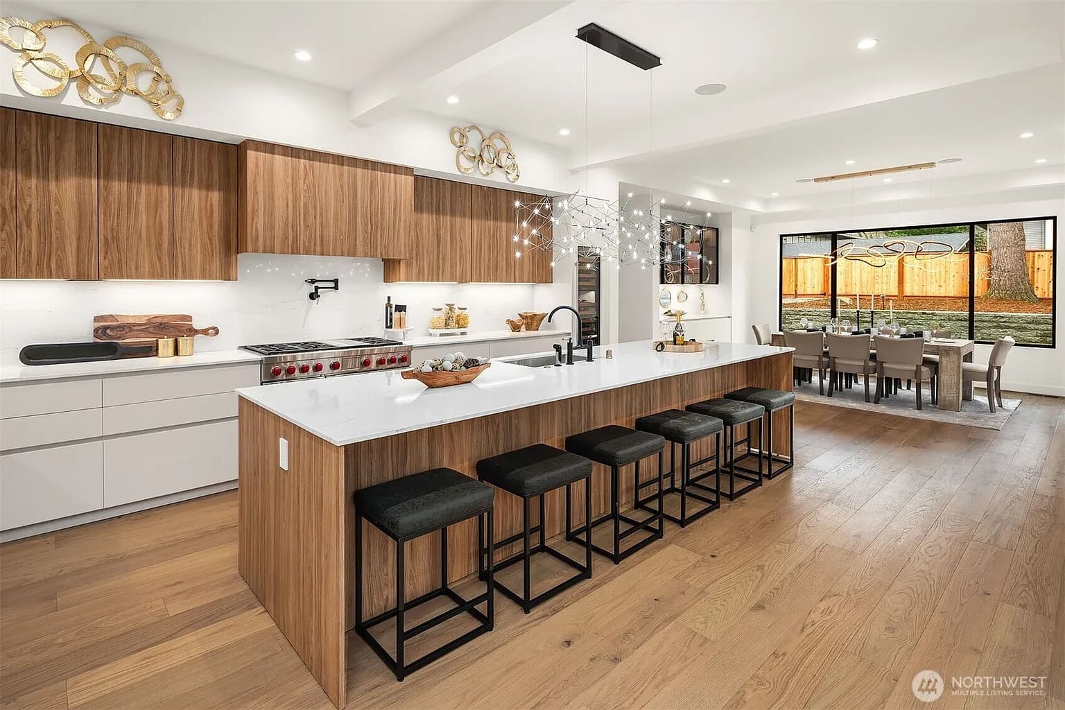 Modern kitchen with large island, wooden cabinets, bar stools, and adjacent dining area with large windows.