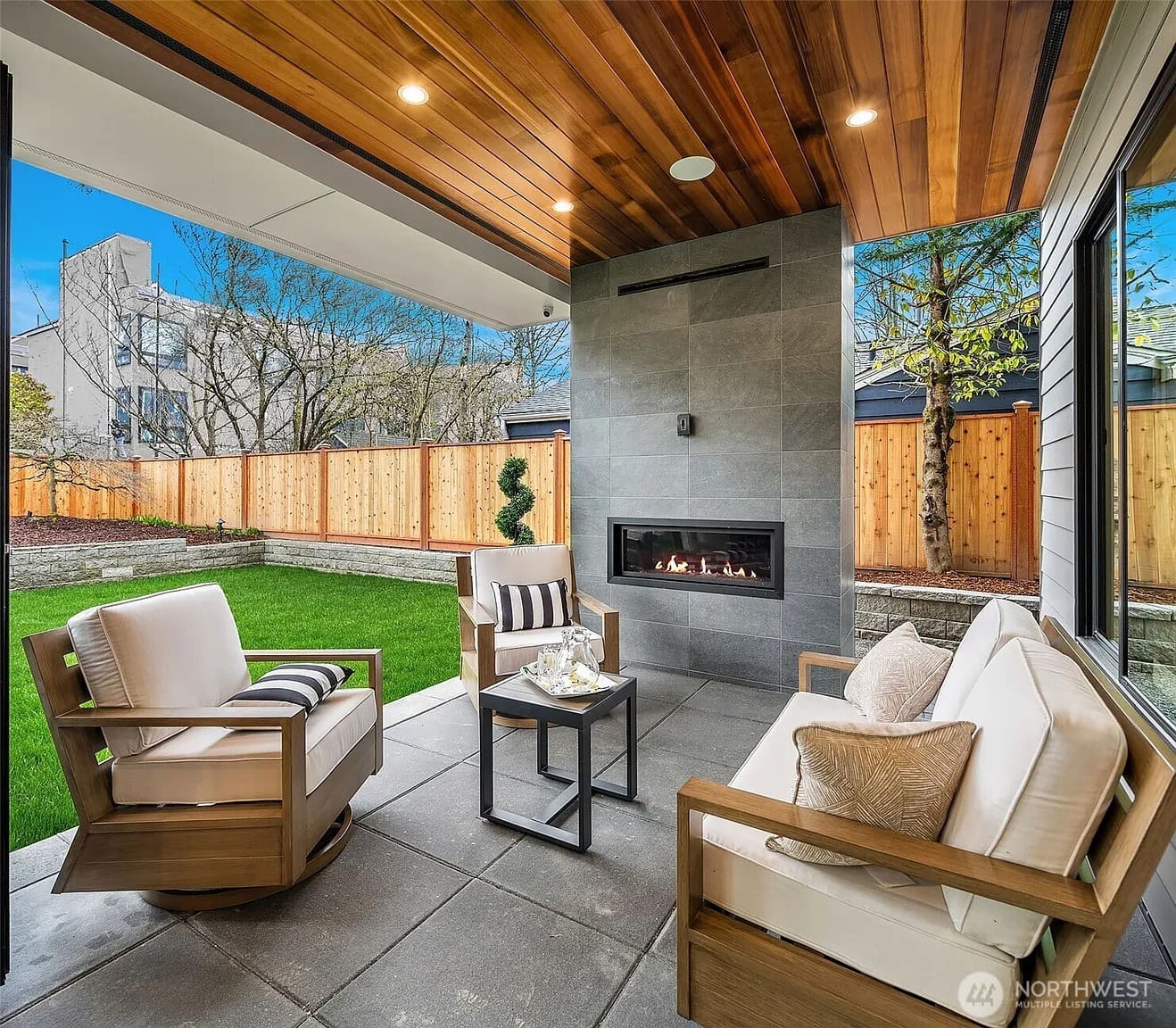 Covered patio with modern furniture, fireplace, and view of a fenced backyard and trees.