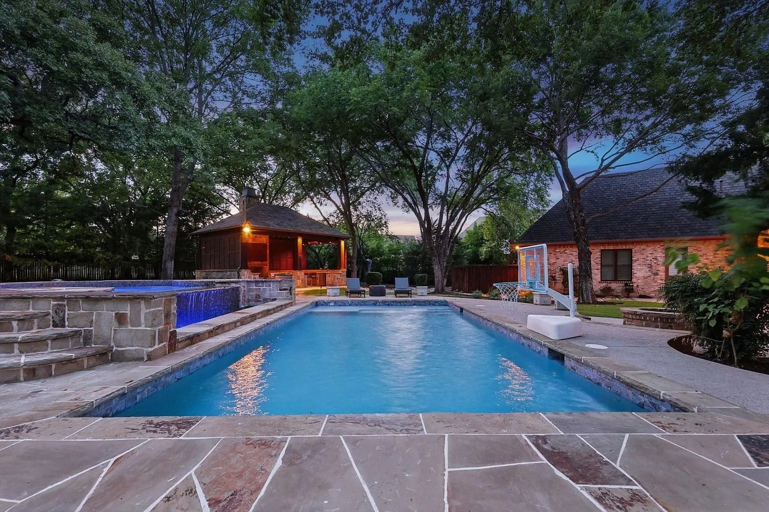 Backyard with a swimming pool, hot tub, patio, and trees at dusk.