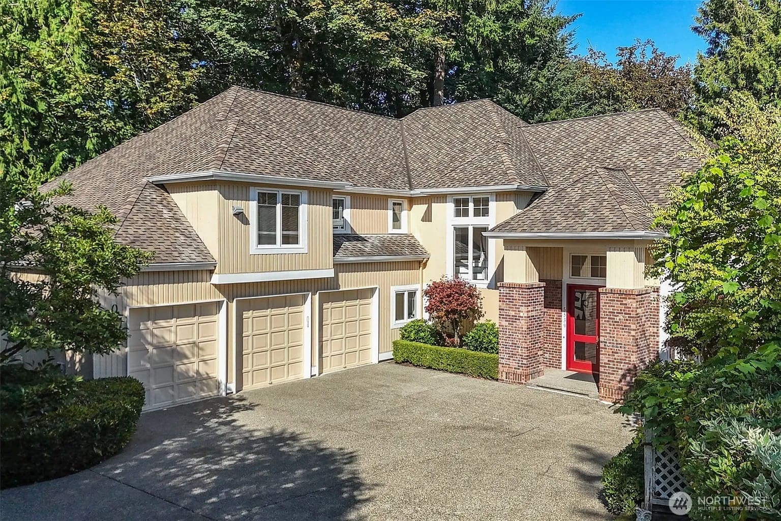 Two-story beige house with a three-car garage, red front door, and surrounded by trees.