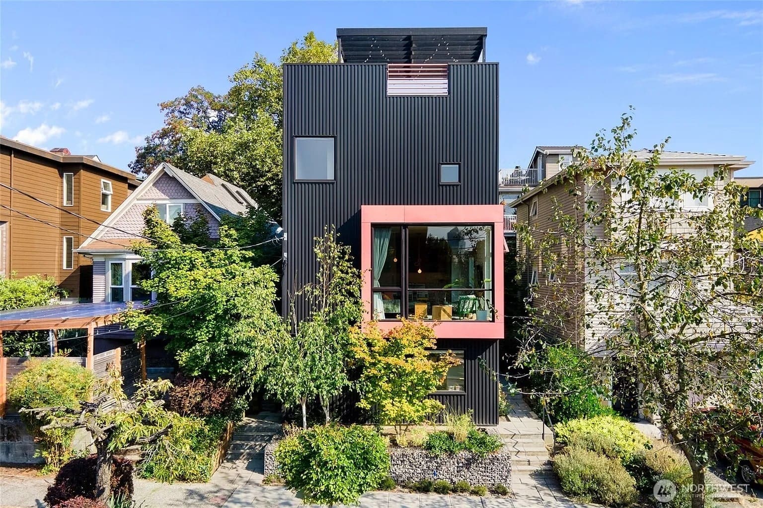 Modern black house with large windows, surrounded by greenery, between traditional homes.