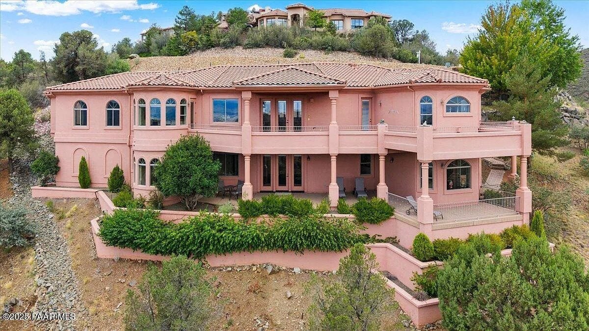 Large pink stucco house with multiple balconies, surrounded by trees on a hillside.