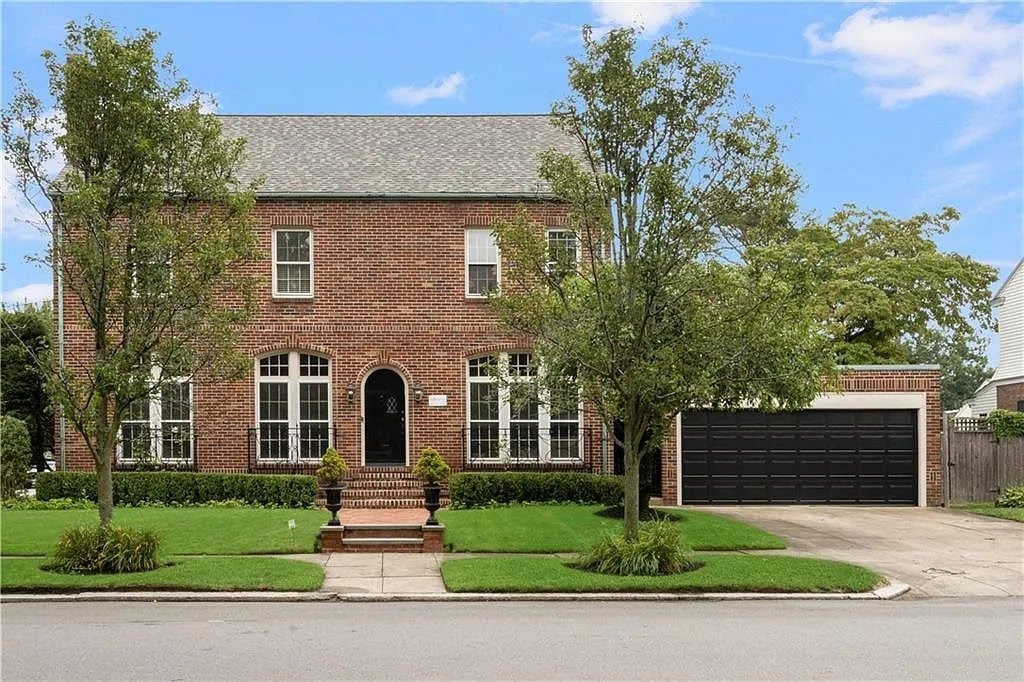 Two-story brick house with a manicured lawn, trees, and a black garage door.
