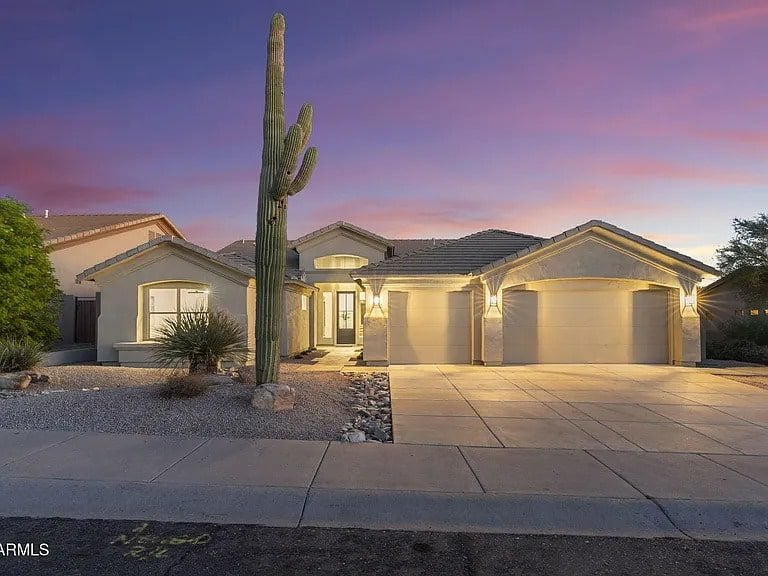 Single-story house with a three-car garage, desert landscaping, and a large cactus at sunset.