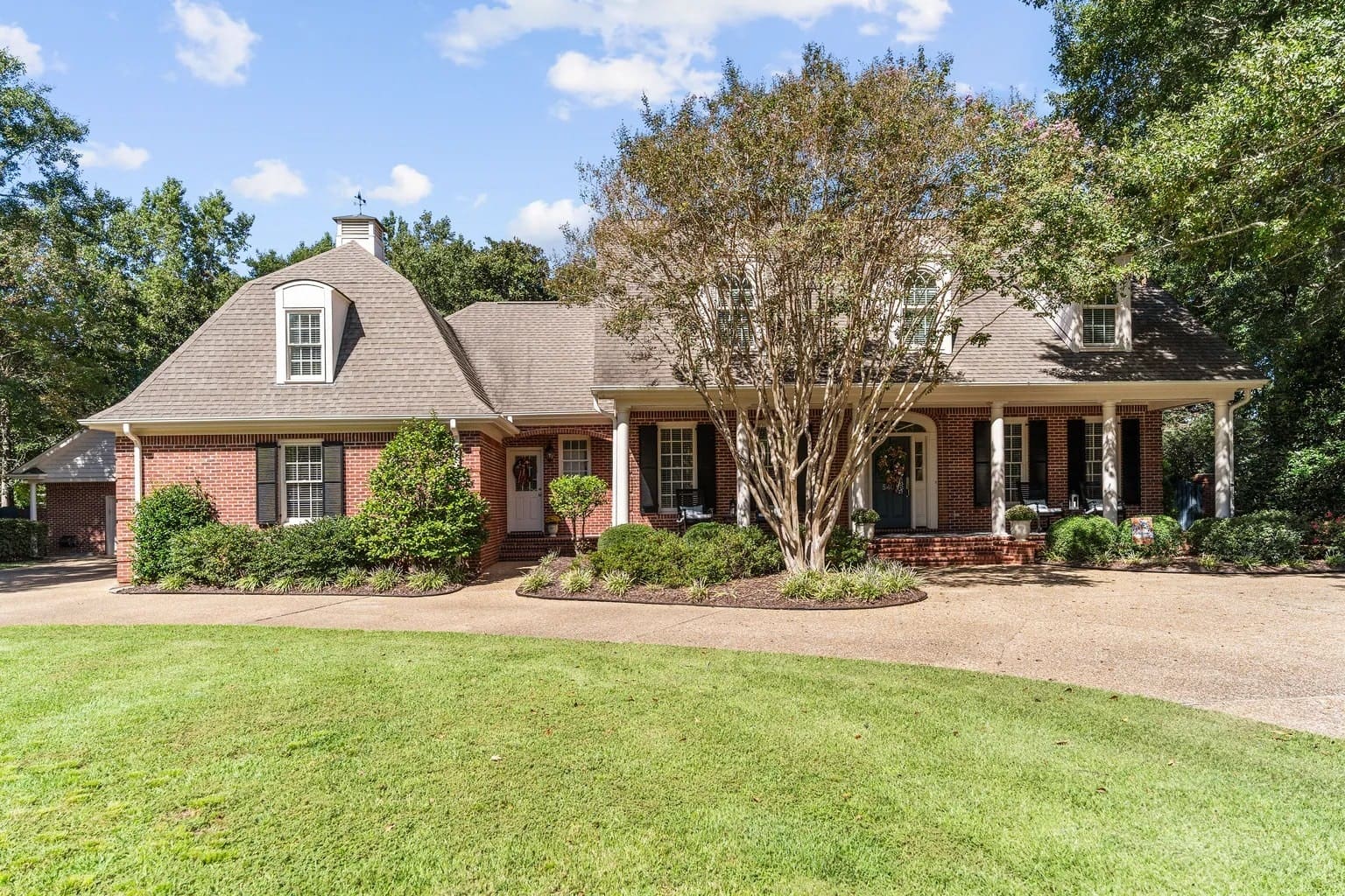 Brick house with a large front yard, trees, and a circular driveway under a blue sky.