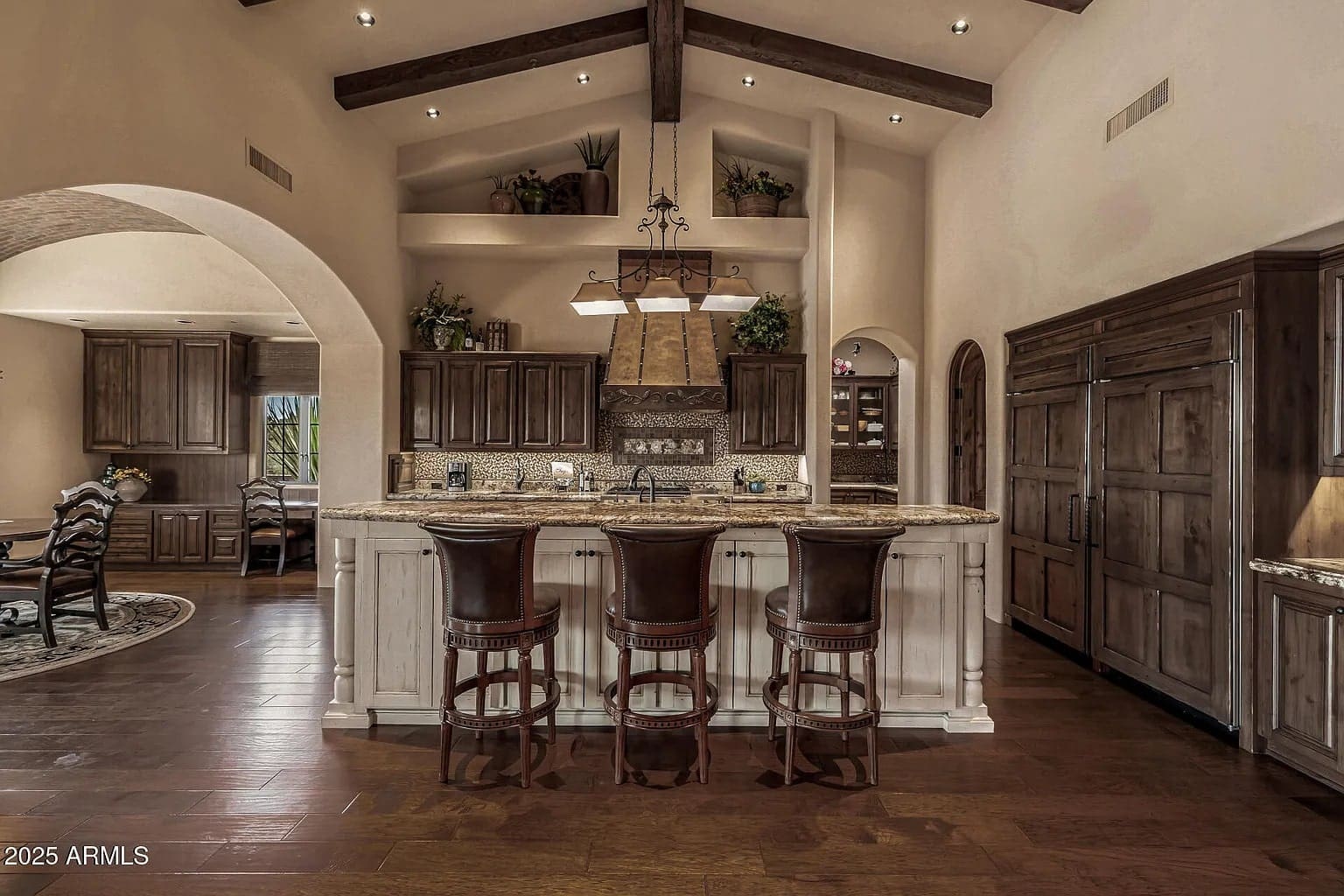 Rustic kitchen with wooden beams, bar counter, three stools, and built-in wooden cabinets.