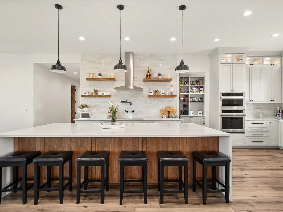Modern kitchen with large island, bar stools, pendant lights, and open shelving on tiled wall.