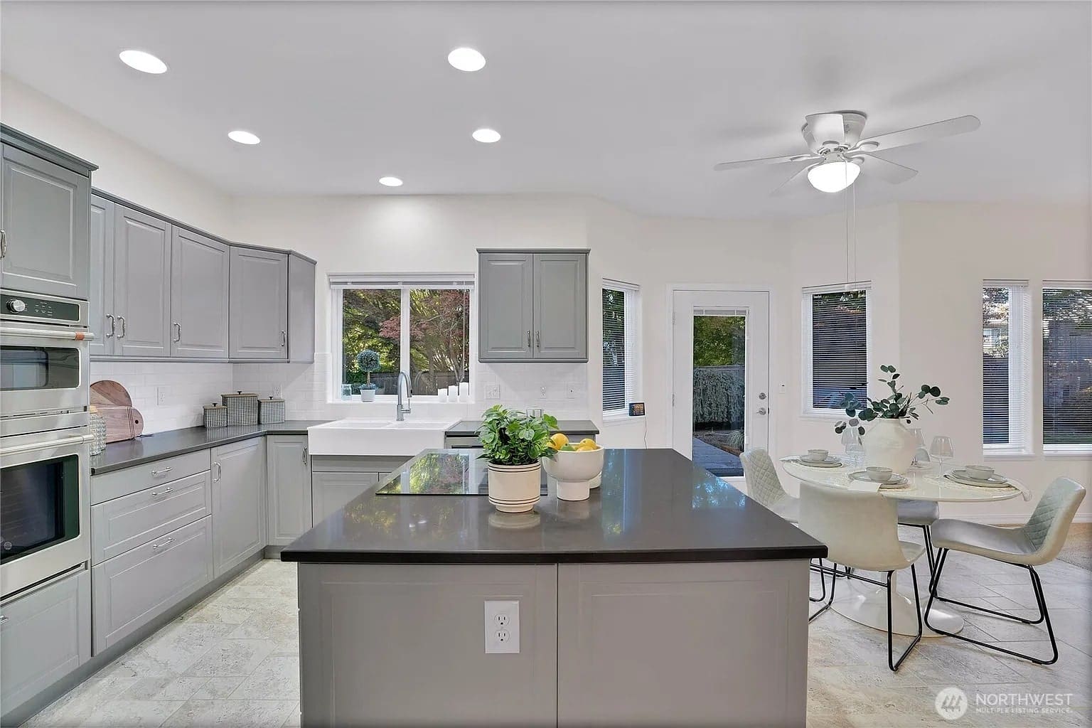 Modern kitchen with gray cabinets, island, and dining area. Large windows and ceiling fan present.