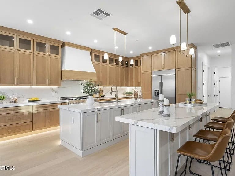 Modern kitchen with wooden cabinets, large island, marble countertops, and bar stools. Bright lighting.