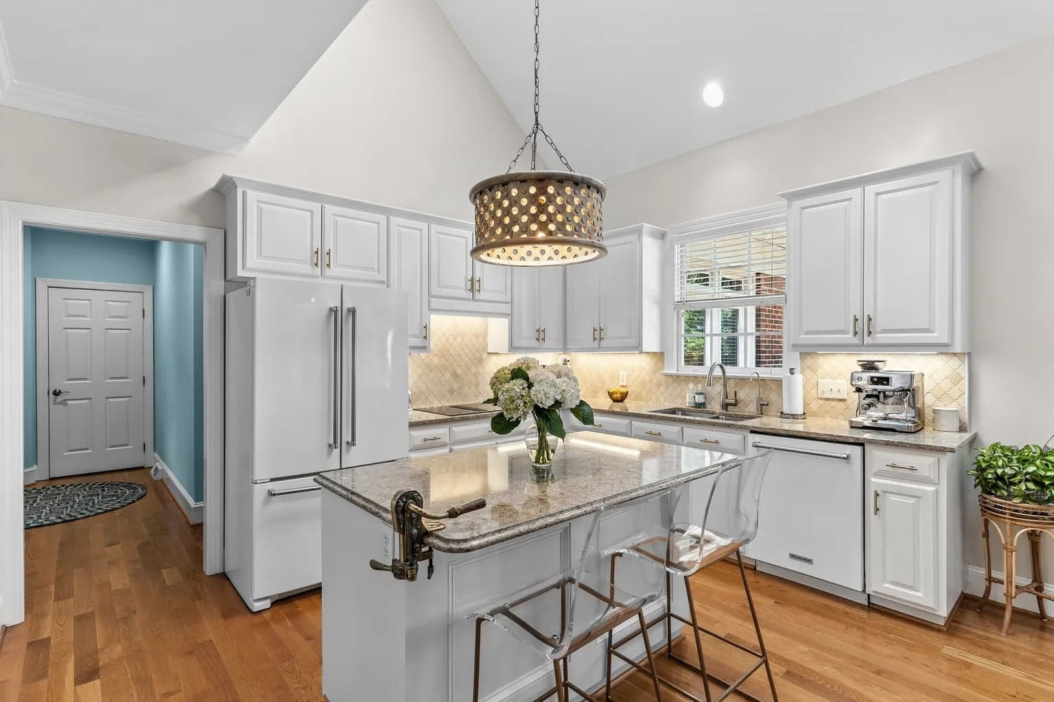 Modern kitchen with white cabinets, island, pendant light, and wooden floor. Doorway leads to another room.