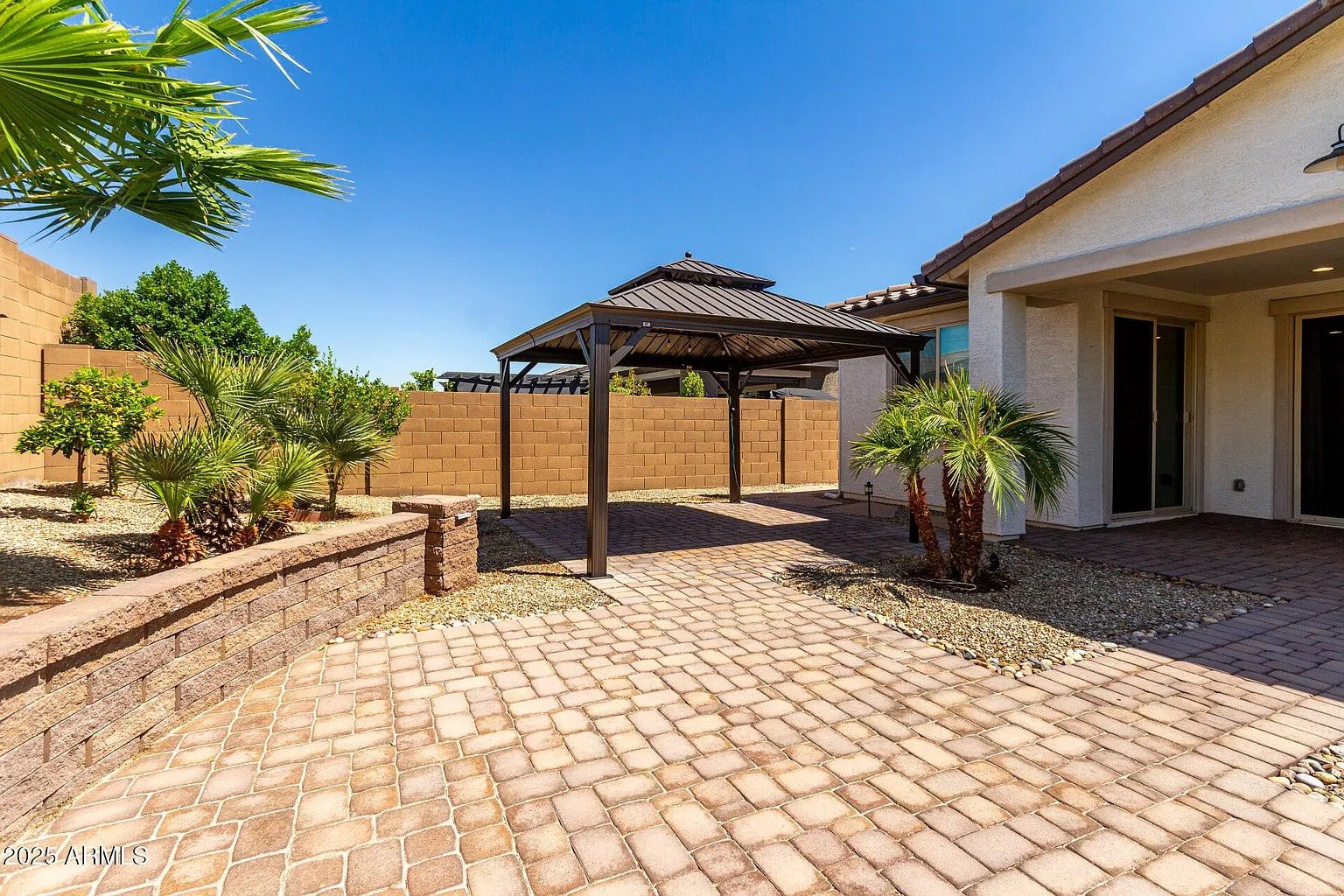Backyard with a paved patio, gazebo, palm trees, and a brick wall under a clear blue sky.