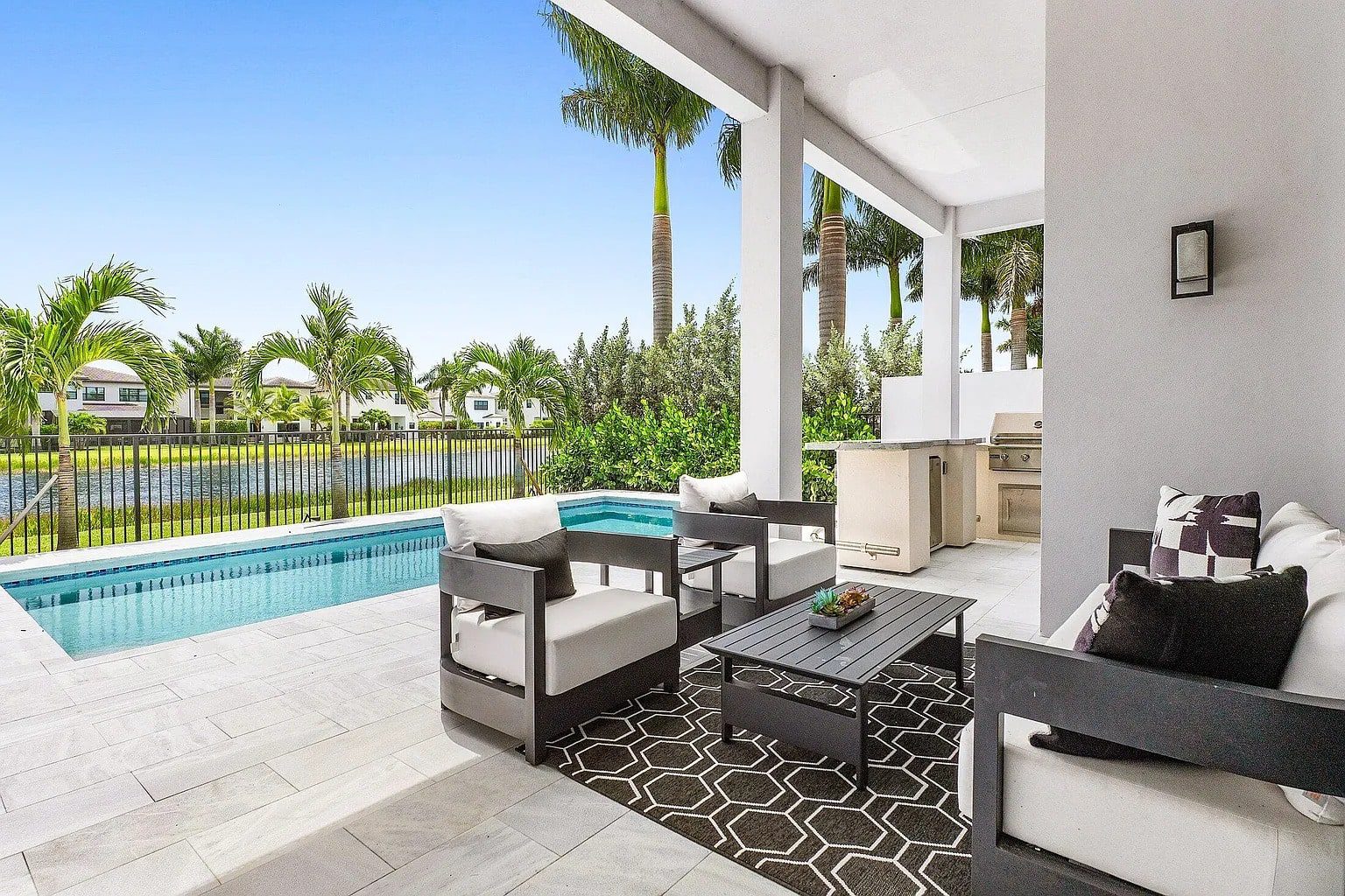 Modern patio with pool, outdoor seating, grill, and view of palm trees and houses.