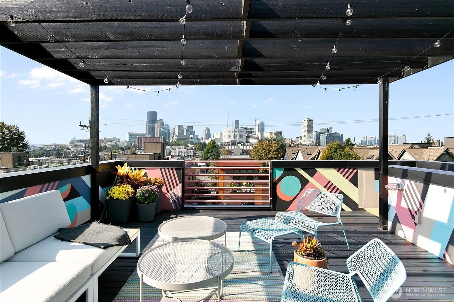 Rooftop patio with colorful furniture, city skyline view, and string lights under a pergola.